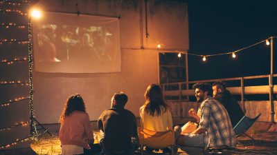 Group of young friends watching a movie on a building rooftop terrace, eating popcorn, drinking beer and having fun